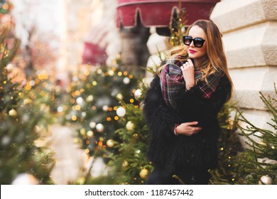 Outdoors Lifestyle Fashion Portrait Of Stunning Blonde Girl. Smiling, Walking On The Christmas City. Going Shopping. Trendsetter. Wearing Stylish Black Fur Coat, Scarf And Sunglasses. Festive Mood