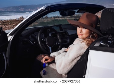 Outdoors Lifestyle Fashion Portrait Of Stunning Young Woman Driving Convertible. Driving Towards The Ocean. Girl Travelling Behind The Wheel. Wearing Stylish Sheepskin Coat, Hat. Enjoying Trip.