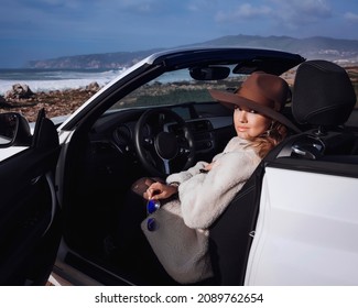 Outdoors Lifestyle Fashion Portrait Of Stunning Young Woman Driving Convertible. Driving Towards The Ocean. Girl Travelling Behind The Wheel. Wearing Stylish Sheepskin Coat, Hat. Enjoying Trip.