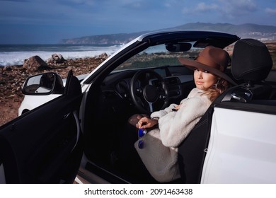 Outdoors Lifestyle Fashion Portrait Of Stunning Young Woman Driving Convertible. Driving Towards The Ocean. Girl Travelling Behind The Wheel. Wearing Stylish Sheepskin Coat, Hat. Enjoying Trip.