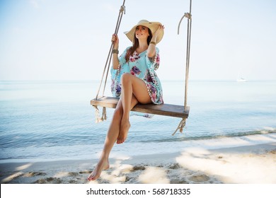 Outdoors lifestyle fashion portrait stunning young girl enjoying on swing on the tropical island. In the background the sea. Wearing stylish blue dress, straw hat and bracelets. Straight long hair