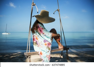 Outdoors lifestyle fashion portrait stunning girl sitting on swing on the beach. Sea background. Wearing stylish blue dress, bracelets and straw hat. Straight long hair. Enjoying life in paradise