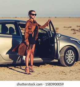 Outdoors Lifestyle Image Of Stunning Young Woman Standing Near The Car. Cyprus. Wearing Stylish Beige Cloak And Sunglasses. Trendsetter. Freedom And Success Concept
