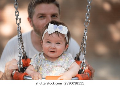 outdoors lifestyle portrait of father playing with beautiful and adorable baby girl smiling cheerful on playground playing on swing at city park wearing a cute ribbon in childhood concept - Shutterstock ID 2330038111