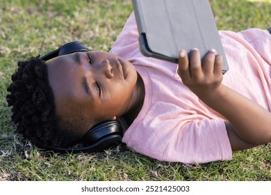 Outdoors, Lying on grass, african american boy wearing headphones using tablet for schoolwork. Learning, technology, studying, outdoors, education, childhood - Powered by Shutterstock