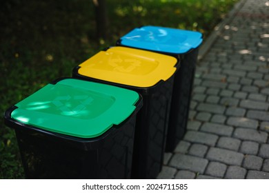 Outdoors in the park is recycling bins different colours. Tree closed plastic bins. Green, yeallow and blue