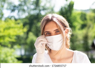 Outdoors portrait of beautiful young woman wearing cotton white mask and medical/surgical gloves on sunny day. She is adjusting mask. Nature/green trees in the background. - Powered by Shutterstock