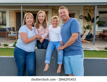 Outdoors Portrait Of A Happy Family Of Four Smiling In Front Of New Dream Home Or Vacation Rental House. Mom, Dad, And Children Boy And Girl, Embracing And Having Fun Together Enjoying Holiday Villa.