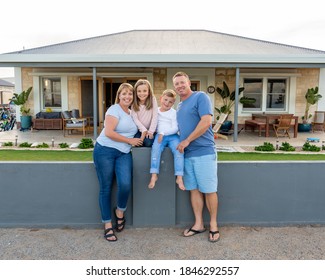 Outdoors Portrait Of A Happy Family Of Four Smiling In Front Of New Dream Home Or Vacation Rental House. Mom, Dad, And Children Boy And Girl, Embracing And Having Fun Together Enjoying Holiday Villa.