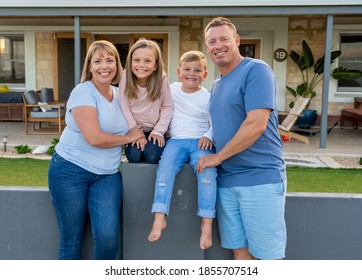 Outdoors Portrait Of A Happy Family Of Four Smiling In Front Of New Dream Home Or Vacation Rental House. Mom, Dad, And Children Boy And Girl, Embracing And Having Fun Together Enjoying Holiday Villa.