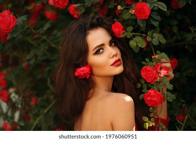 Outdoors Portrait Of Natural Beautiful Woman In Red Roses. Sensual Female With Rose Flowers In Hair. Sexy Brunette With Makeup Over Sunset Blossom Park. Passion And Expression. Spanish Flamenco Style.