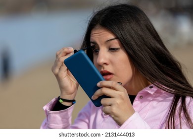 Outdoors portrait of young beautiful teen  using smartphone as mirror while do her makeup standing on the banks of the lake 