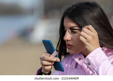 Outdoors portrait of young beautiful teen  using smartphone as mirror while do her makeup standing on the banks of the lake 