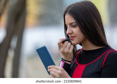 Outdoors portrait of young beautiful teen  using smartphone as mirror while do her makeup