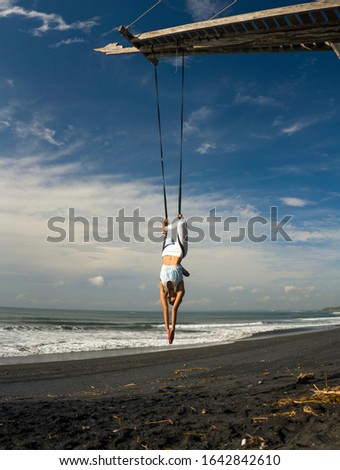 outdoors portrait of young happy and athletic Asian Indonesian woman doing aero yoga beach workout training body balance and relaxation hanging from swing rope above the sea in healthy lifestyle
