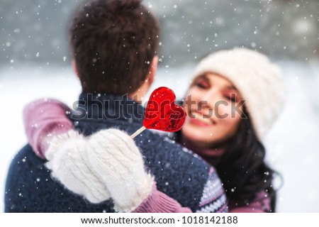 Outdoors romantic portrait of happy hugging couple in love. Girl smiling and holding in her hand red lollipop in shape of heart. Wearing knitted hat, mittens and sweaters. Love concept. Valentine day