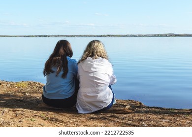 Outdoors, Two Women Dressed In A Blue Hoodie And A White Jacket With Flowing Hair Are Sitting On The Shore Of A Reservoir, Looking From The Back And Looking Ahead At The Horizon, Copy Space.