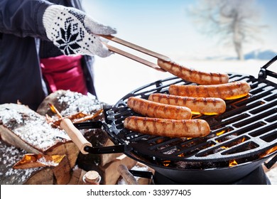 Outdoors winter barbecue party with a person wearing knitted woollen gloves cooking sausages over hot coals in a BBQ, close up view