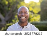 Outdoors, young African American man smiling in sunlit garden. Lush greenery background creating a serene atmosphere, with vibrant plants and flowers adding to peaceful scene, unaltered