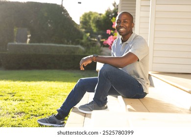 Outdoors, young African American man sitting on porch steps, smiling at camera. Sunlit garden and house exterior in background, creating warm and inviting atmosphere, unaltered - Powered by Shutterstock