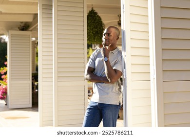 Outdoors, young African American man standing on porch, looking thoughtful. Bright, sunny day with flowers and greenery around house, creating peaceful atmosphere, unaltered - Powered by Shutterstock