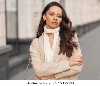 Outdor Portrait. Elegant Woman In Beige Jacket With White Scarf Walking City Street Holding Leather Handbag. Pretty Female Model With Long Wavy Brunette Hair And Perfect Makeup