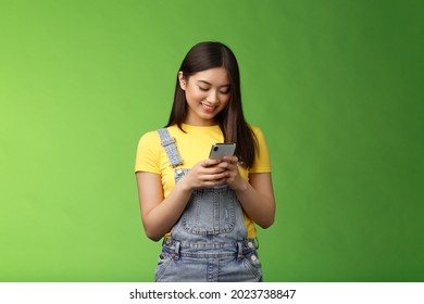 Outgoing cute asian brunette woman scroll online shop, purchase summer tour internet, hold smartphone, look amused telephone screen, use app, edit photos post social media, stand green background - Powered by Shutterstock