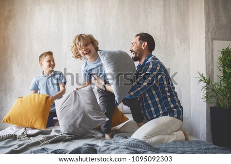 Outgoing dad having fun with two little sons while sitting on cozy bed. Entertainment concept