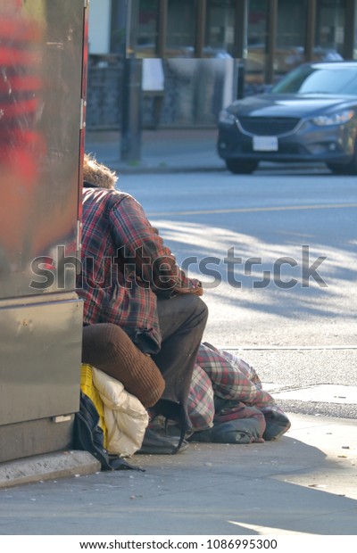 The outline\
of a homeless person in tattered clothing sitting on a street\
corner with his worldly belongings.\

