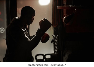 Outline of young African American male boxer in punch gloves standing in gym and hitting sandbag hanging on wall during sports training - Powered by Shutterstock