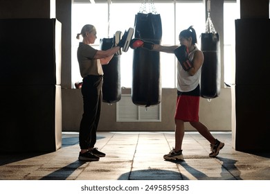 Outlines of boxing trainer holding defense punching pads while standing in front of young female boxer during kick training - Powered by Shutterstock