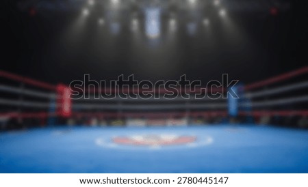 An out-of-focus image of a boxing ring, showcasing the ropes, corners, and floor, illuminated by overhead lights, creating a sense of anticipation and space.