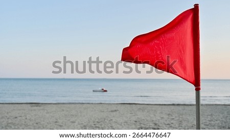 Out-of-focus red triangular warning flag in the foreground, with a sharp focus on the calm but potentially dangerous sea in the background. A pedal boat floats near the horizon.	
