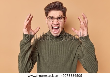 Outraged man gestures angrily raises shakes hands and screams loudly expresses fury has fight with wife wants to get divorce dressed in casual jumper isolated over beige background. Feel my fury