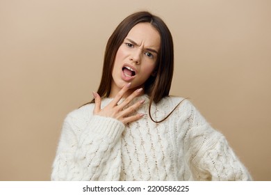 The Outraged Woman Stands And Looks Straight At The Light Brown Background In A White Sweater, Frowning Menacingly With Her Eyebrows Raised To Her Face With Outstretched Fingers