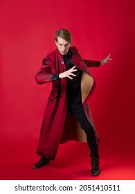 An Outrageous Young Man In A Daring Red Coat In A Vintage Noir Style, Epic Poses And Makes A Hand Gesture. Photo On A Red Background