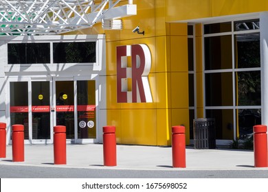 The Outside Entrance To An Emergency Department Of A Hospital. The Exterior Wall Of The Building Is Bright Yellow Metal Composite Panels With Windows And White Metal Overhang And Red ER Letters.