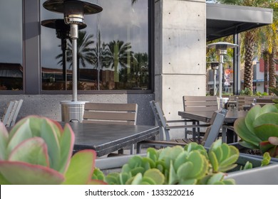 An Outside Seating Area Of A Restaurant With Green Plants In The Foreground. The Patio Area Has A Number Of Heaters.