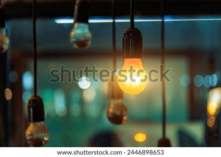 Outstanding Concept. One glowing light bulb among dark unlit lamps. Concept of unique idea, creativity, and leadership. Vintage Edison light bulb glowing warmly with beautiful colorful bokeh background at night. Photo. Illuminated incandescent lightbulb hanging on a wire close up with blurry lights background