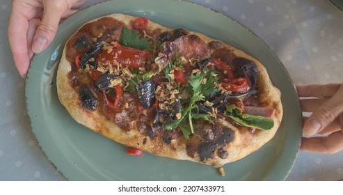 Oval Mini Pizza Lies On A Plate. Top View, Hands Rotate The Plate. Appetizing Portion Of Italian Food For One Person. Burnt Dough, Tomatoes, Salami.