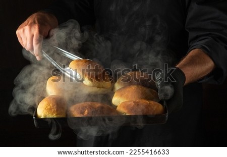 Oven baked buns on a hot baking sheet in the baker hand in glove. The cook takes a bun with a culinary tool.