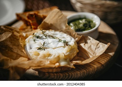 Oven baked camembert cheese with rosemary and pesto sauce on baking paper. Homemade grilled brie with thyme on wooden plate. Rustic style table setting. Cottage core, local vacation