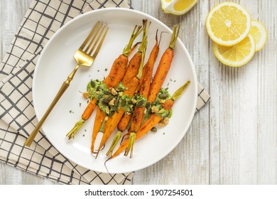 Oven baked whole carrots in lemon juice Easter side dish; round white plate on wooden board with carrots green mayonaise dressing, walnuts, pumpkin seeds and herbs; vegetarian meal