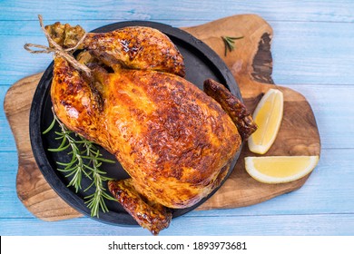 Oven Baked Whole Chicken On A Black Ceramic Plate On A Kitchen Wooden Board With A Slice Of Lemon And Baked Garlic And A Sprig Of Rosemary.Grilled Chicken.