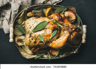 Oven roasted whole chicken with onion, apples and sage in serving tray over dark stone background, top view, selective focus. Celebration food concept
