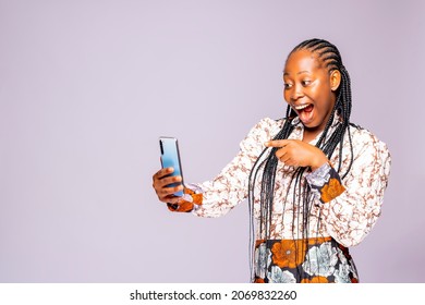 Over Excited Black African Female Hipster Pointing At Her Phone With Her Mouth Widely Open. Young Lady African Using Cellphone Texting In Social Media Standing On A White Studio Background