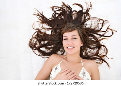 Over Head Close Up Beauty Portrait Of An Attractive Hispanic Young Woman Laying Down With Her Brunette Hair Curling Around Her, Joyfully Smiling. Exterior Lifestyle.