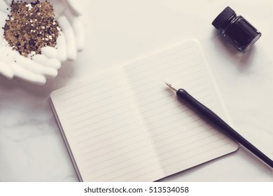 Over head flat lay view of a white and gold chic desktop. Open notebook, pen and ink, gold office supplies and  open hands holding glitter.