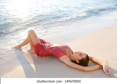 Over Head Side View Of An Attractive Young Woman Relaxing On The Shore Of A White Sand Beach, Wearing A Pink Dress And Bathing In The Waves While Laying Down Being Thoughtful.