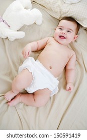 Over Head View Of A Beautiful Baby Girl Laying Down On A Bed At Home With Her Soft Toy Friend, Joyful And Smiling, And Wearing A White Nappy.